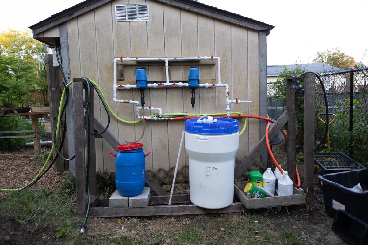 Two blue fertilizer injectors on a white PVC manifold on the shed wall, with a blue drum and a white bucket below