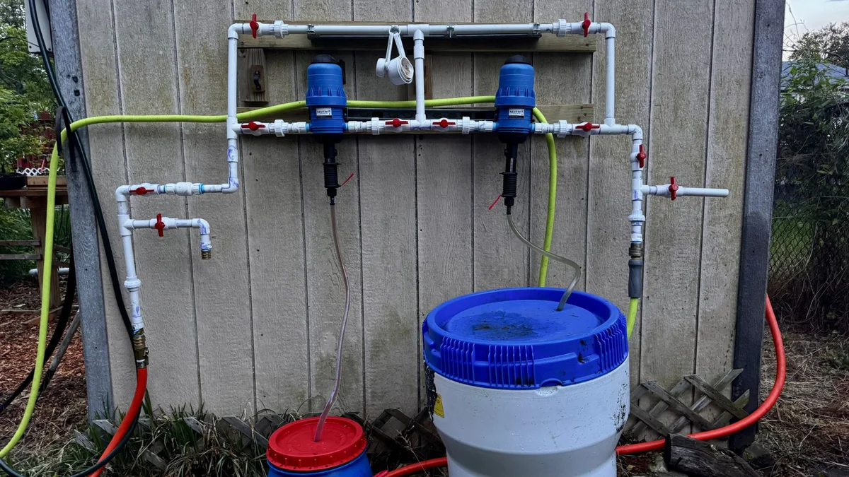 Two blue NEWTRY injectors on a white PVC manifold with red-handled valves, mounted on the shed wall, each drawing from a container below